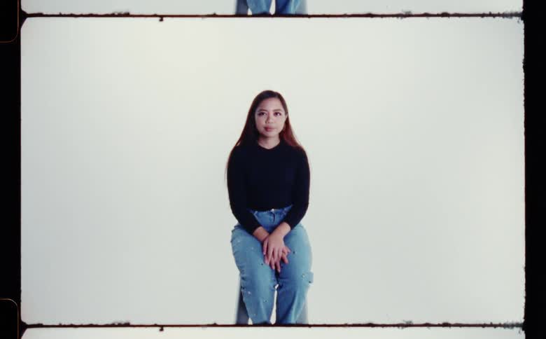 A woman wearing a black shirt and jeans sitting on a stool with her hands in her lap against a white backdrop