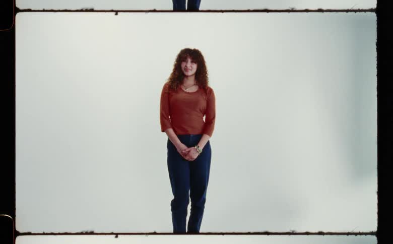 Woman with curly hair brown shirt and dark blue pants smile at camera in front of white screen backdrop in photographic studio