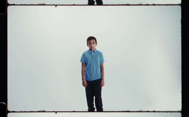 A boy wearing a blue collared shirt and black pants standing with arms at his sides in front of a white backdrop