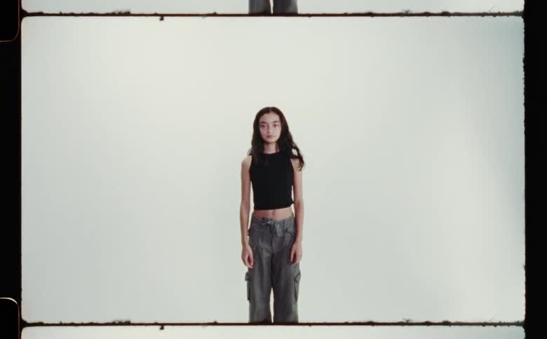 A teenage girl wearing a black belly shirt and grey pants standing with arms at her sides against a white backdrop