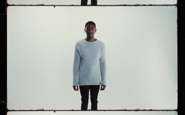 A man in a sweater and black pants standing with his eyes closed and then open against a white backdrop