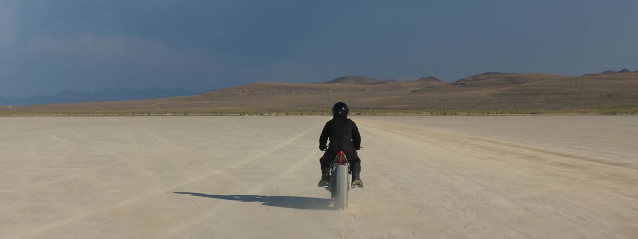 View from behind of a person wearing a helmet riding a motorcycle through he desert on a sunny day with blue sky