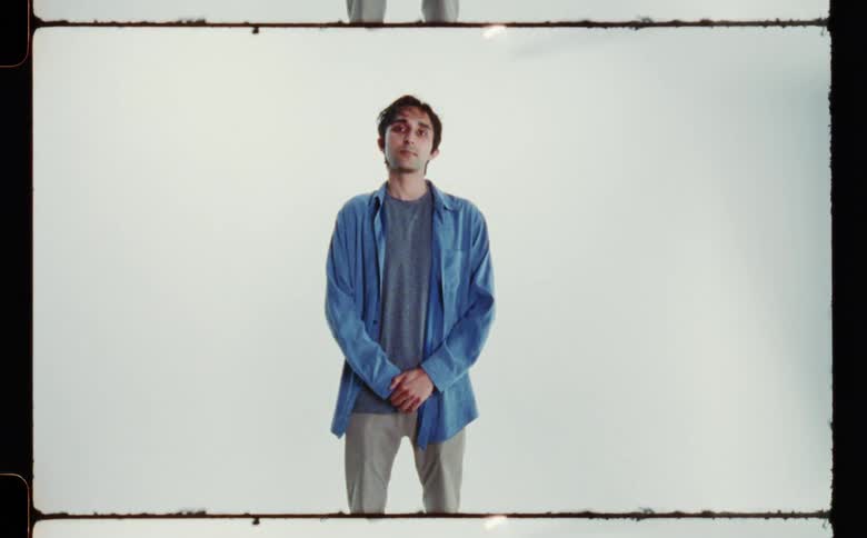 A man wearing a blue overshirt and tan pants standing with his hands in front of him against a white backdrop