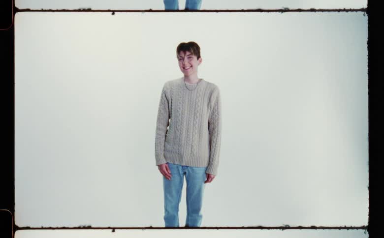 An androgynous teen smiles nervously while standing posed in front of a blank screen