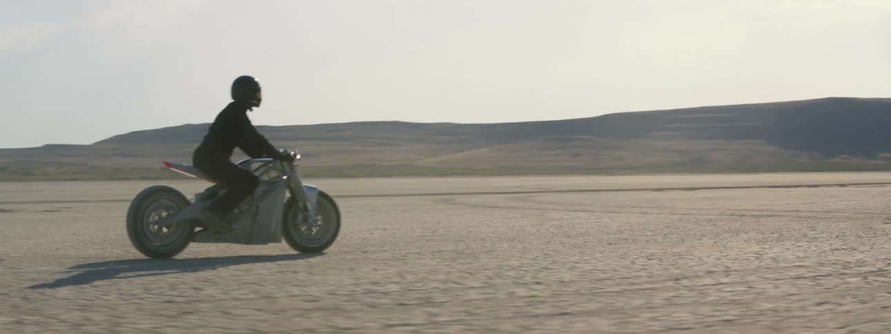 A motorcycle travels across the desert as the rider is wearing a black jacket and helmet and there are mountains in the distance