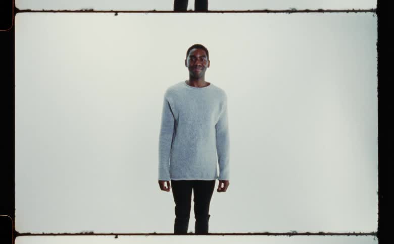 A man wearing a sweater and black pants standing and smiling against a white backdrop