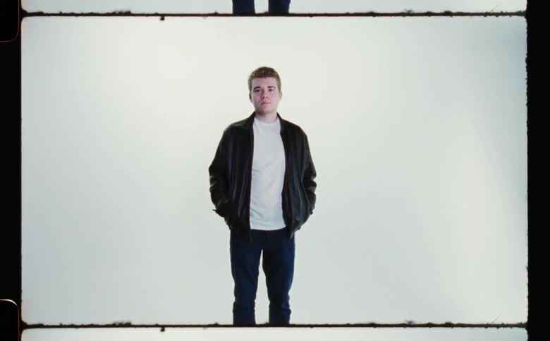 A man wearing blue jeans and a white shirt with a dark jacket is standing with his hands in his jacket pockets as he looks ahead