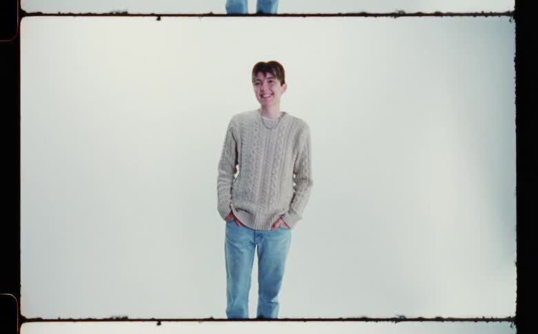 A teenage boy wearing a sweater and blue jeans is standing with his hands in his pockets smiling and laughing