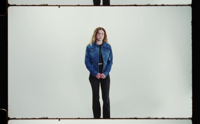A teenage girl with long brown hair is standing and looking ahead wearing black jeans and a blue denim jacket