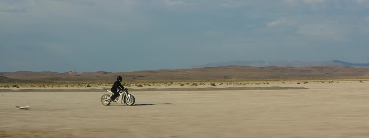 A motorcycle travels across the desert as the rider is wearing a black jacket and helmet and there are mountains in the distance