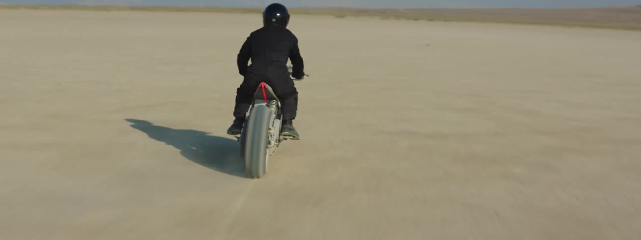 A motorcycle travels across the desert as the rider is wearing a black jacket and helmet and there are mountains in the distance