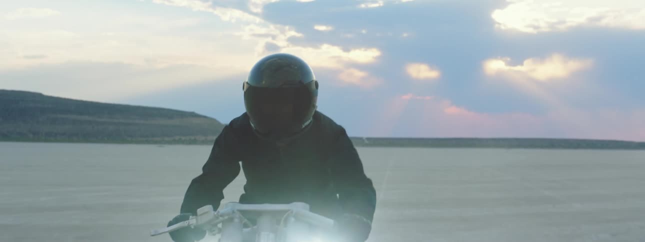 Motorcyclist rides motorbike with its headlights on across desert with mountains in the background and a red tinted horizon in the distance