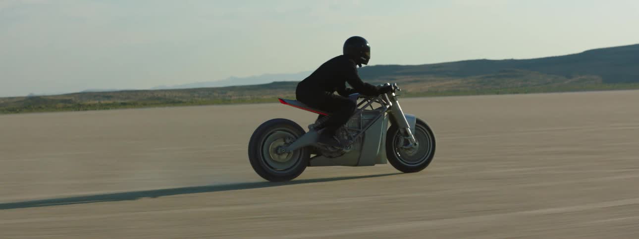 A motorcycle travels across the desert as the rider is wearing a black jacket and helmet and there are mountains in the distance
