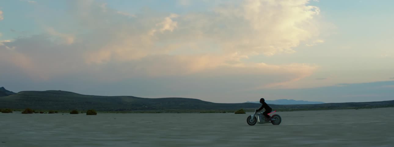 A motorcycle travels across the desert as the rider is wearing a black jacket and helmet and there are mountains in the distance under a cloudy sky