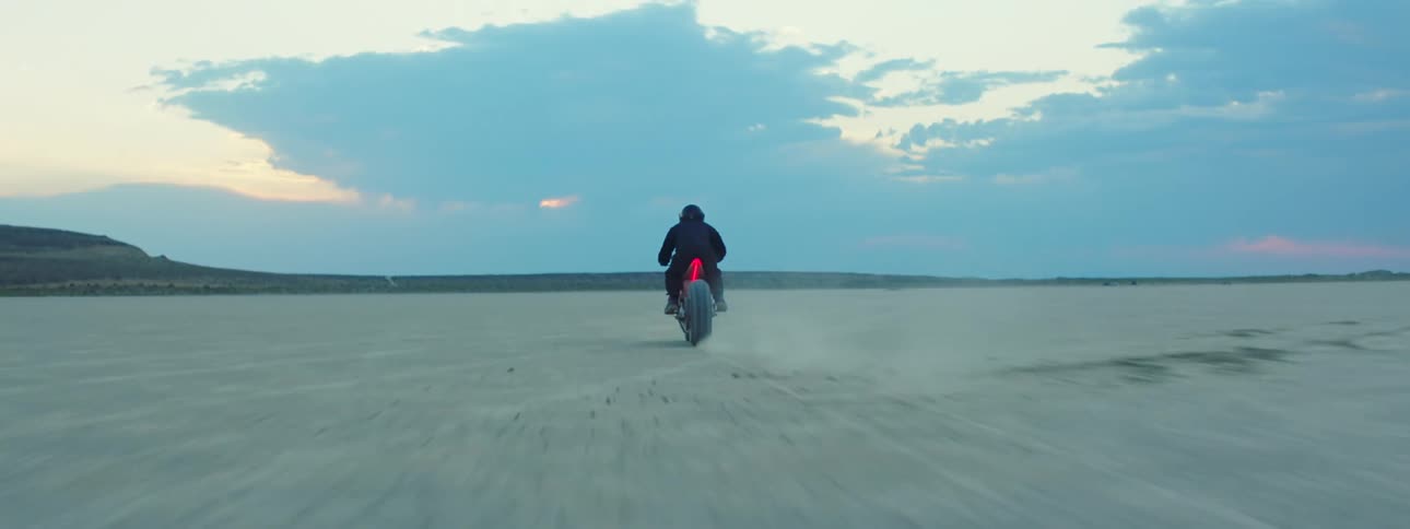 A motorcycle speeds across the desert leaving a trail of dust as the rider is wearing a black jacket and helmet and there are mountains in the distance
