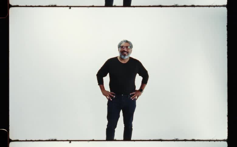 A man wearing a black shirt and dark pants is standing with his hands on hips and a smile on his face