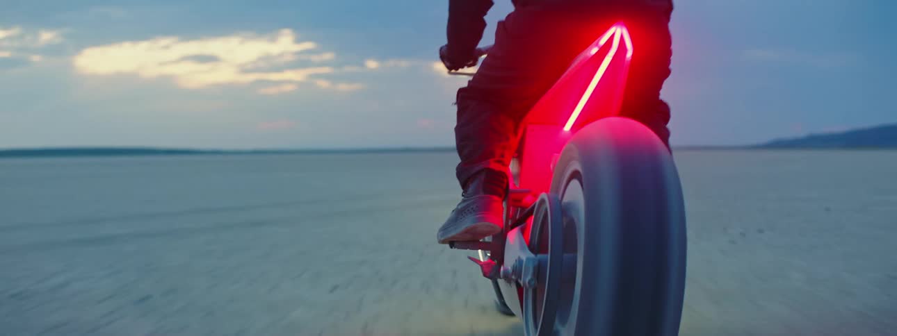 Motorcyclist rides motorcycle lit by red lighting across arid plain with mountains in distance