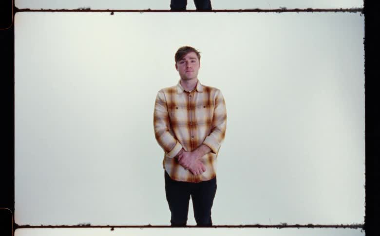 Man with brown plaid shirt and black jeans poses for the camera in front of white screen backdrop in photography studio