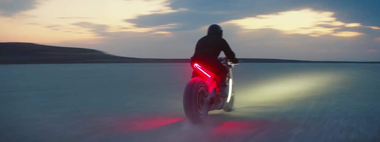A motorcycle travels across the desert with the headlight on and a red light glowing under the seat as the rider is wearing a black jacket and helmet