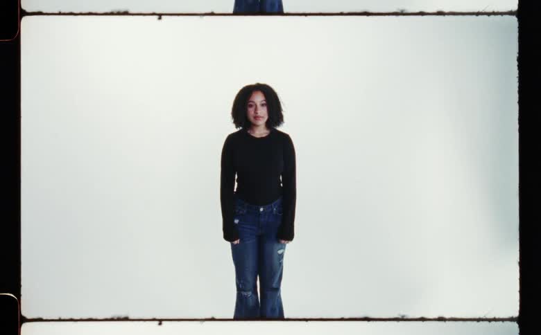 Woman with curly hair black shirt and ripped jeans poses for the camera in front of white screen backdrop in photography studio