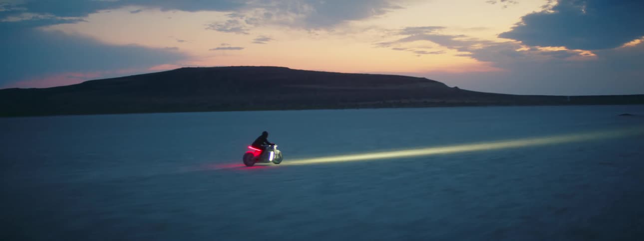 A motorcycle travels across the desert with the headlight on and a red light glowing under the seat as the rider is wearing a black jacket and helmet