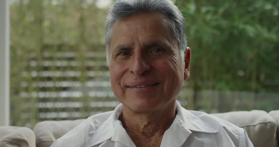 A man is sitting on a couch near a window with a smile on his face as he turns his head and looks forward