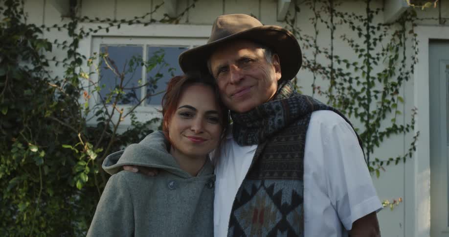 Man with hat gray scarf and white shirt and woman wearing gray hoodie smiles at camera in front of building
