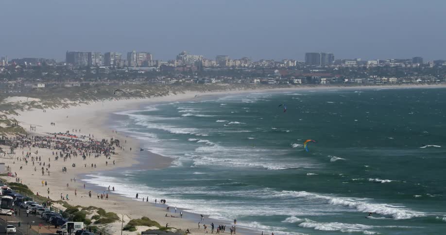The beach is filled with people and kitesurfers flying over the water with tall buildings of a city skyline are in the distance