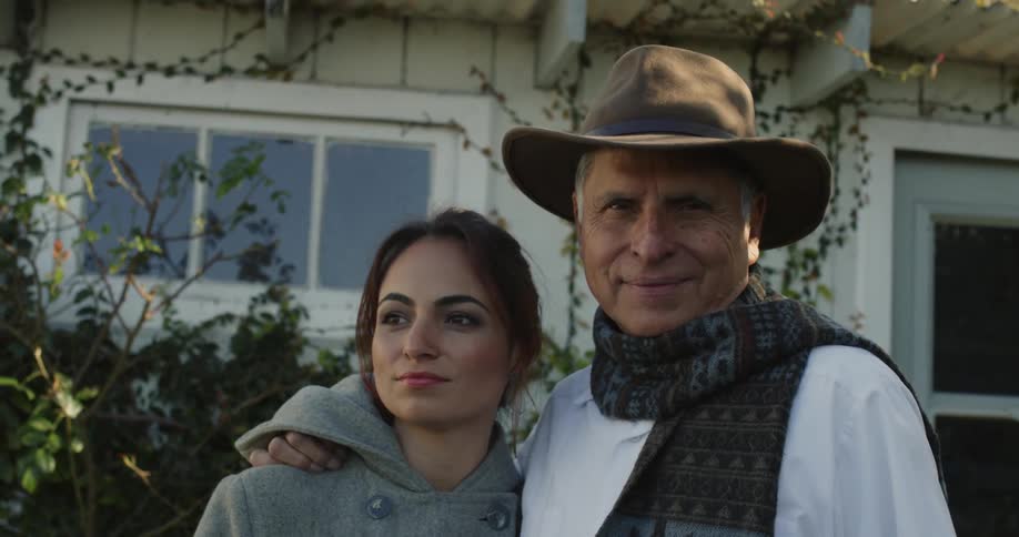 A man wearing a cowboy hat and a scarf around his neck is standing outside of his house with his arm around his daughter