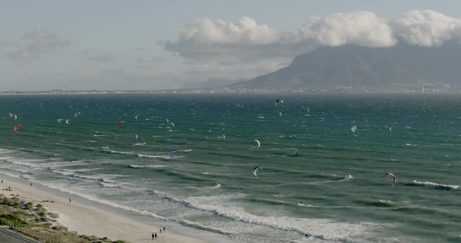 People are Kitesurfing in the waves off a white sand beach with mountains in the distance under a cloudy sky