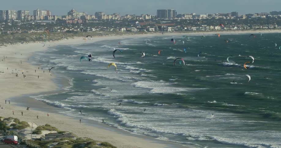Aerial view of kitesurfers kitesurfing in the waves and ocean near a white sand beach with city skyline in the background