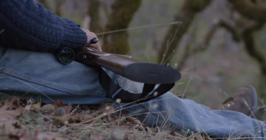 A man is sitting outside on a hill with his hand on his rifle as it lies across his legs