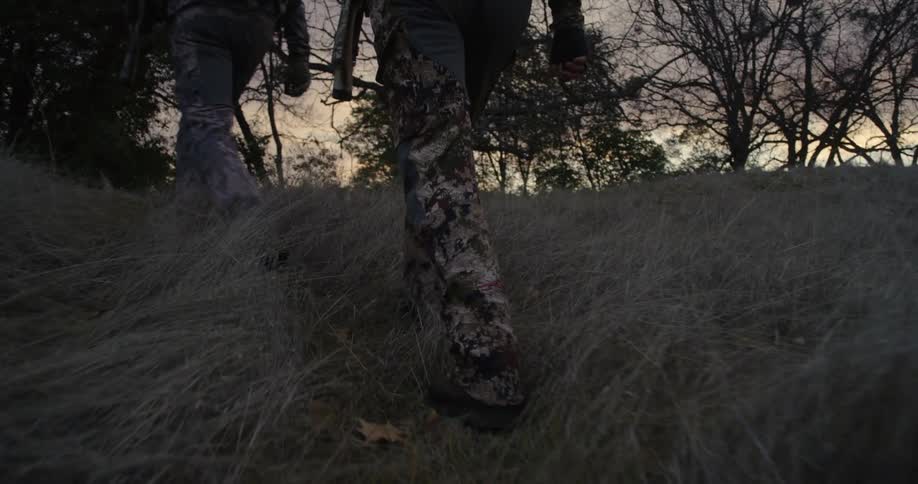 Two hunters wearing camouflage are walking up a hill towards trees