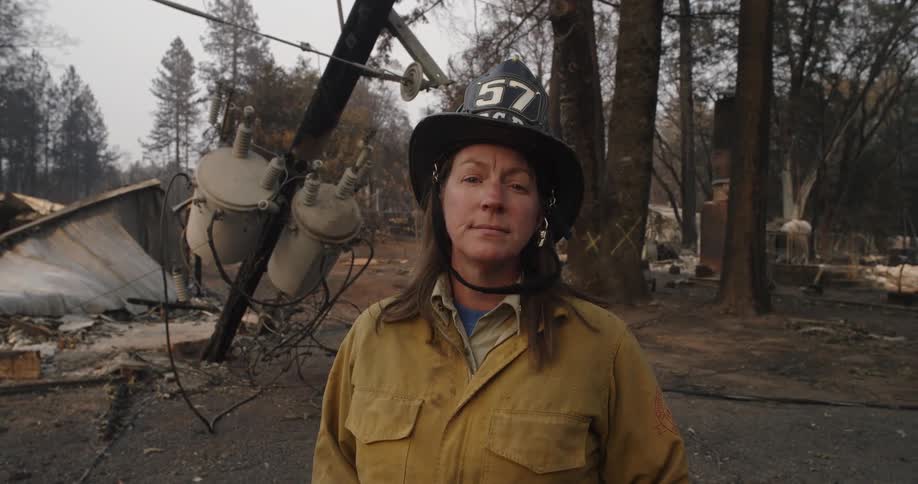 A woman in a fire fighters hat stands in front of a patch of charred trees and land near a powerline that is on the ground and a collapsed building beside it with a somber look on her face
