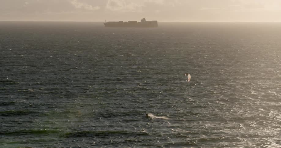 A kitesurfer sails towards a large ship in the open ocean at sunset