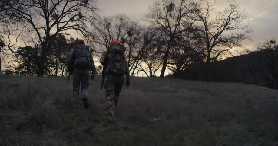 A man and woman wearing camouflage and carrying rifles walk towards barren trees at sunset