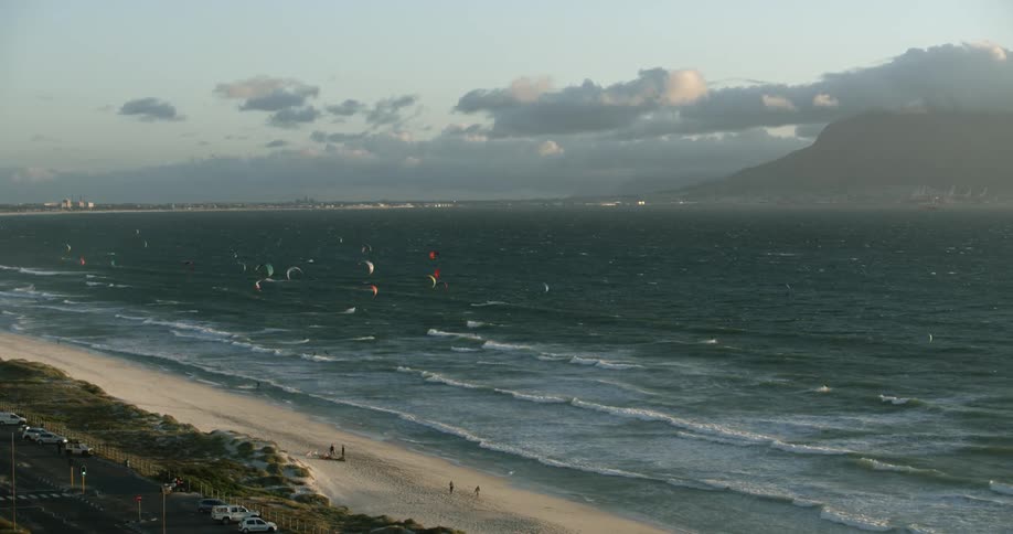 Aerial view of kiteboarders kitesurfing on wavy sea near a white sand beach with Table Mountain in the background