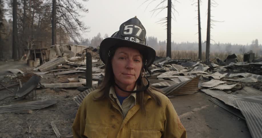 Female government worker wearing helmet and uniform stands near ruined building on edge of woods on foggy day