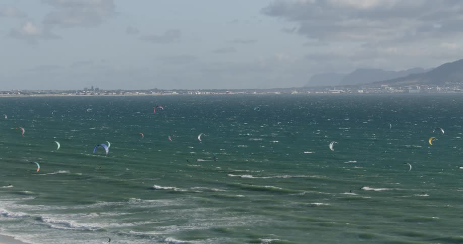 A group of kitesurfers are sailing across the ocean waves near the shoreline with mountains in the distance