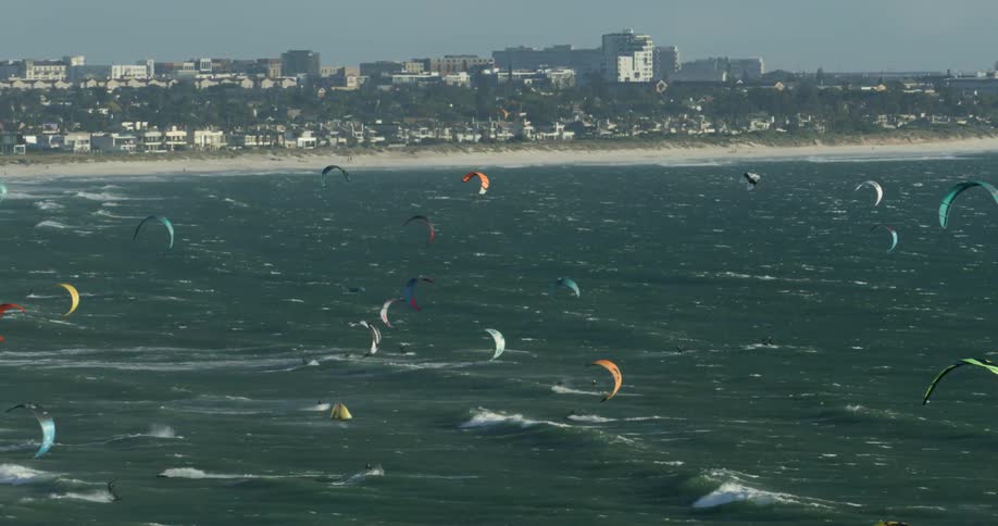 An aerial view of the ocean with many kitesurfers jumping off the waves and landing in the water near a long beach coastline with a large city in the distance