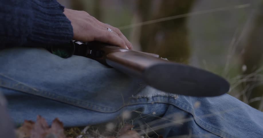 Person sits on ground outdoors with weapon across their legs