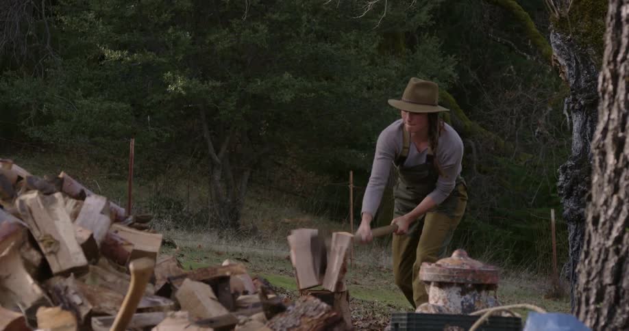Woman with green hat and overralls swinging ax cutting wood