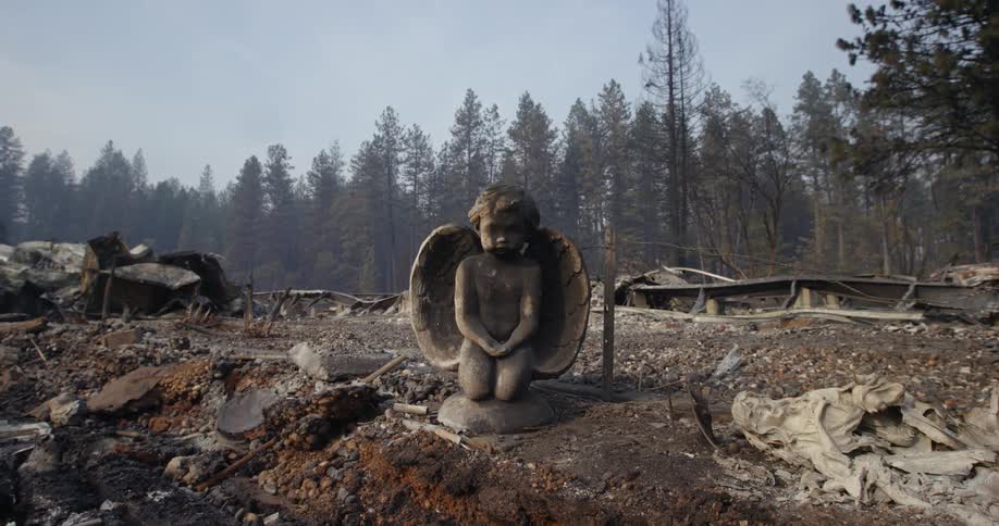 A statue of an angel kneeling with broken discarded statues and fire destruction around it