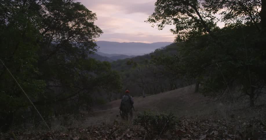 A man and woman hunting and carrying rifles on their backpacks walk down a hill in the evening