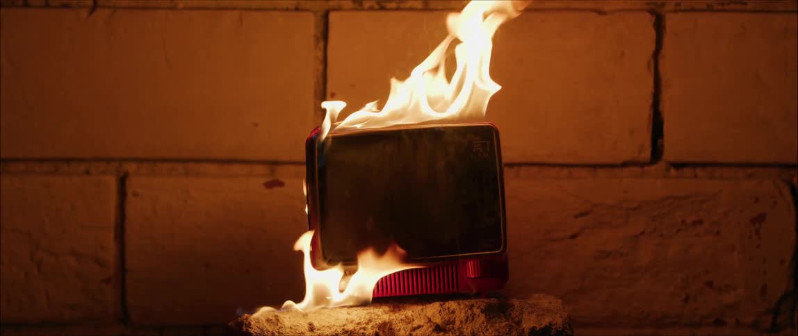 A portable television is in flames as it burns near a brick wall
