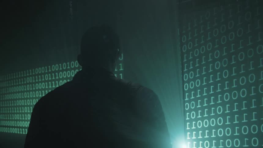 A man stands in front of a screen that is flashing thousands of 1's and 0's