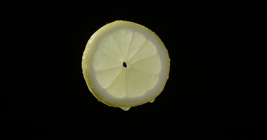 Light shines on a fresh yellow lemon slice with dripping juice turning in front of black background