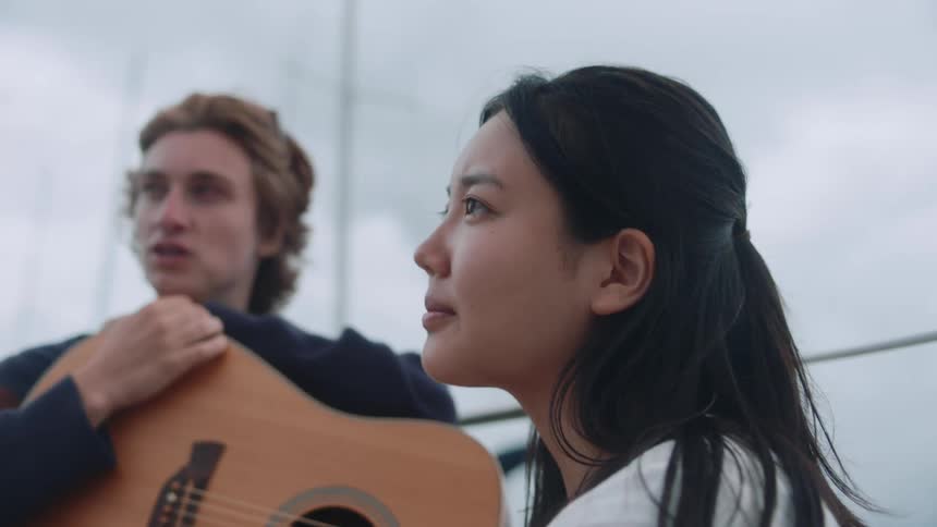 Woman with straight hair white shirt talks to person next to man holding guitar and chewing food on cloudy day