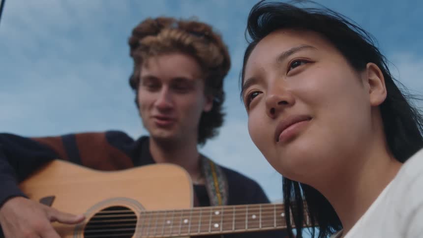 A young woman smiles as the wind blows her hair and a man is playing an acoustic guitar