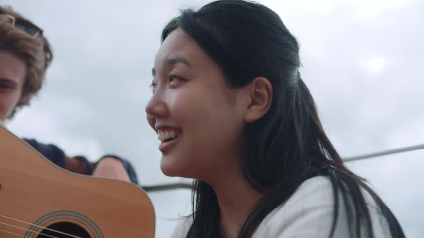 A young woman is smiling and sitting outside next to a young man holding a guitar